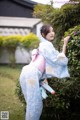 A woman in a blue and pink kimono leaning against a bush.